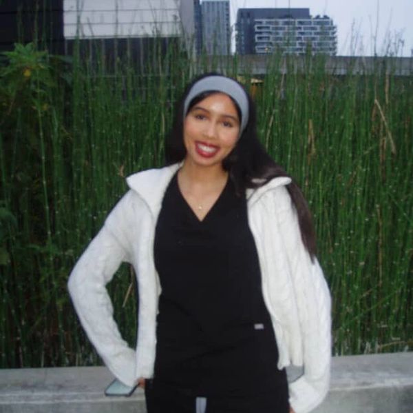 A smiling woman in a white jacket and headband stands outdoors against greenery.