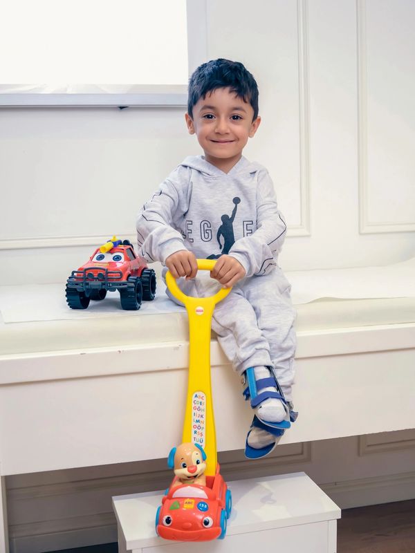 A smiling boy in a gray tracksuit sitting with toys, wearing a foot brace.