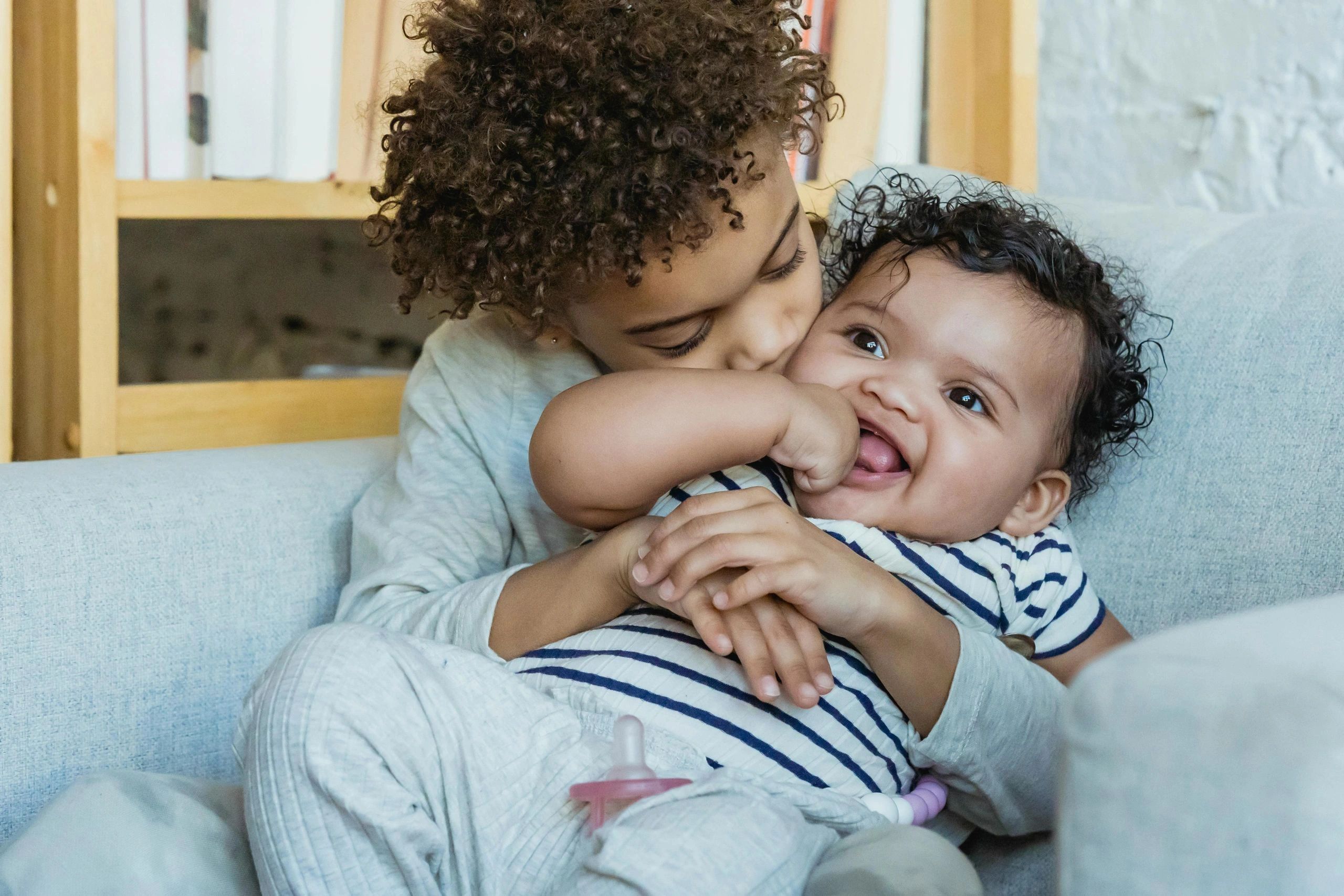 Older sibling affectionately hugs and kisses a happy baby on a couch.