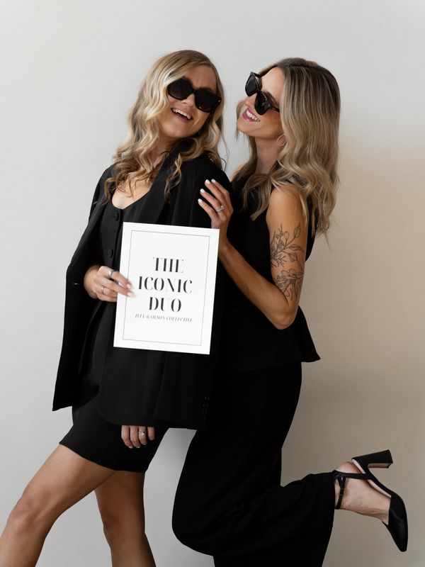 Two stylish women in black pose joyfully, one holding a sign reading 'The Iconic Duo.'