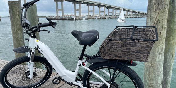 White electric bike with a basket parked by the water and a bridge in the background.