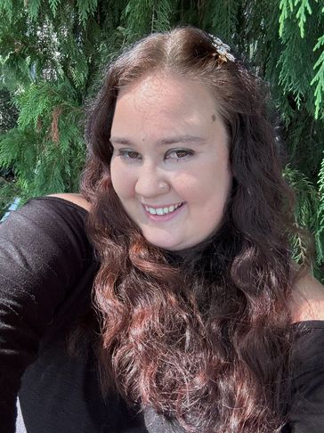 Smiling woman with long wavy hair outdoors by green trees.