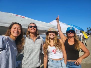 Four women smiling happily at a sunny beach event with tents in the background.