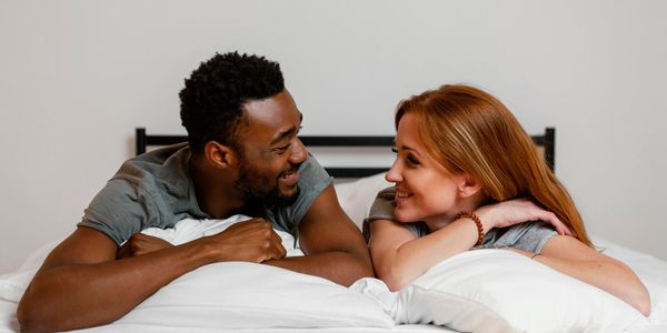 A couple smiling and lying face-to-face on a bed, sharing a warm moment.