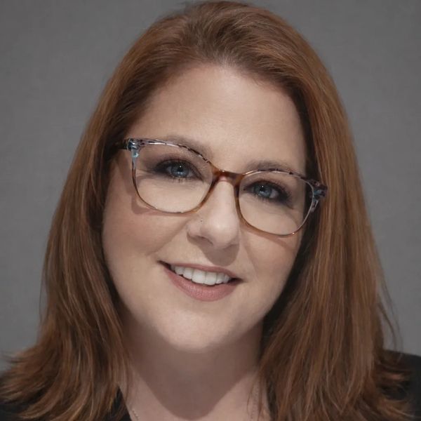 Smiling woman with glasses and red hair against a neutral background.