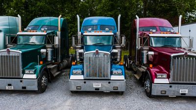 Three colorful semi-trucks parked side by side on a gravel lot.