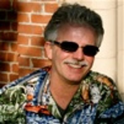 Man in sunglasses and colorful shirt smiles against a brick wall.