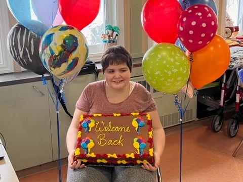 A client of SDS poses between two bunches of balloons while holding a cake that says "Welcome Back".