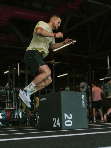 A man jumps onto a plyometric box in a gym, mid-air during a workout.