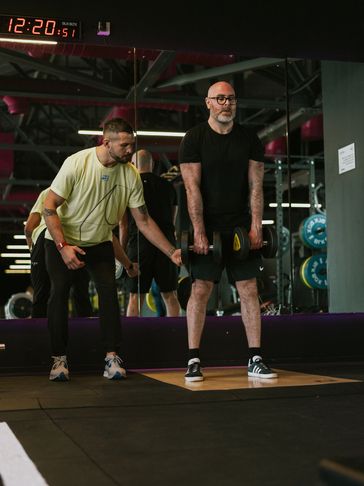 A fitness coach assists a man lifting dumbbells in a gym.