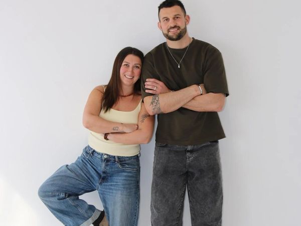 Two people smiling casually against a plain wall, arms crossed.