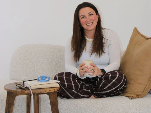 A woman sits cross-legged on a couch, smiling and holding a mug.