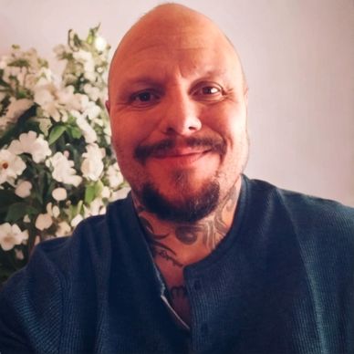 Smiling bald man with a beard and neck tattoos in front of white flowers.