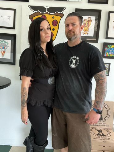 The owners of the shop both stand for a portrait in front of the gallery wall.  