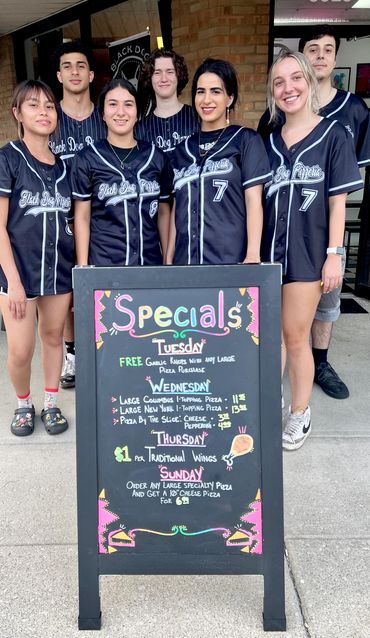 Multiple employees in matching uniforms smiling in front of an advertising sandwich board.  
