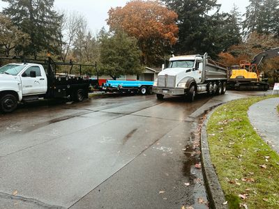 Construction vehicles parked on a wet road near trees and a grassy curb.