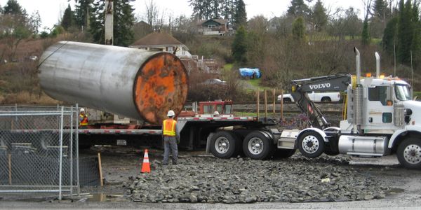Worker supervises heavy cylindrical tank loading onto a flatbed truck at a construction site.