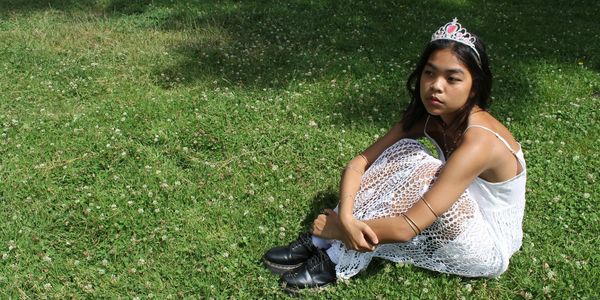 A girl in a white dress and tiara sitting on grass.