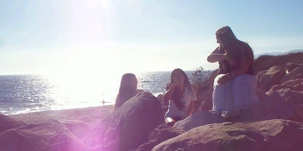 Three people sitting on rocks by the ocean in bright sunlight.