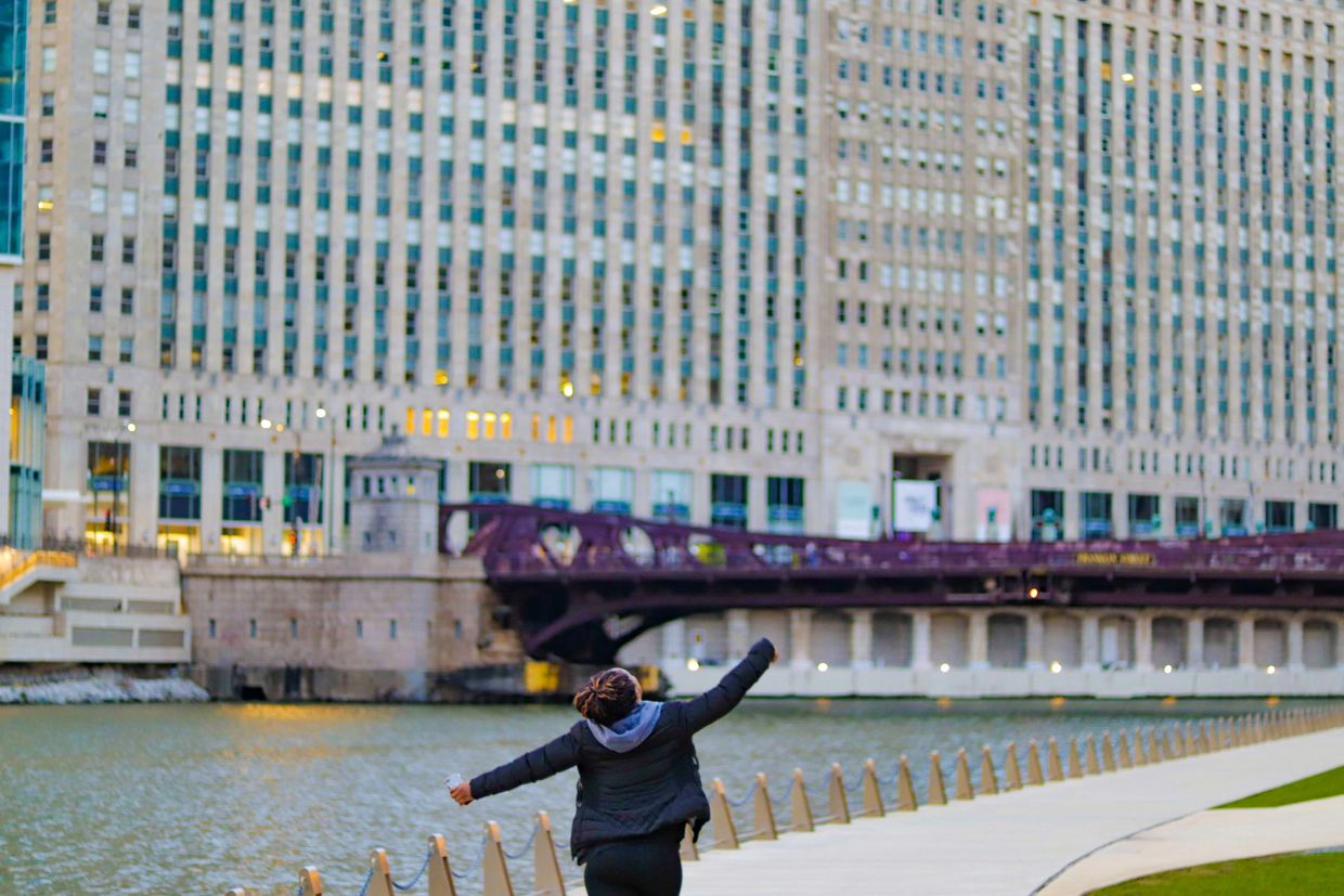 Person joyfully stretches arms near a river with urban buildings in the background.