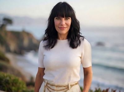 Woman with dark hair stands near the ocean during sunset.