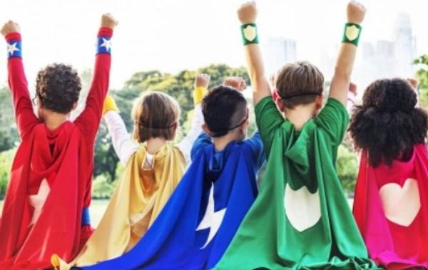 Five children wearing colorful superhero capes with raised fists outdoors.