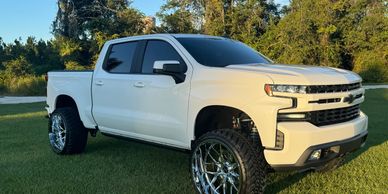 White lifted pickup truck with large chrome wheels parked on grass.
