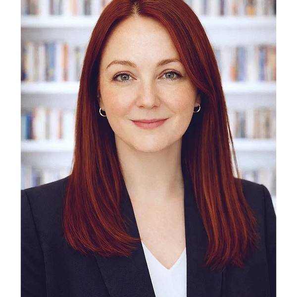 Professional woman with red hair and a confident smile in front of a bookshelf.