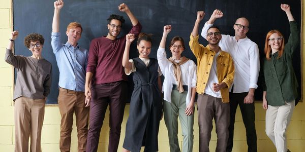 Mixed group of adults smiling in front of a chalkboard with fists raised in accomplishment.