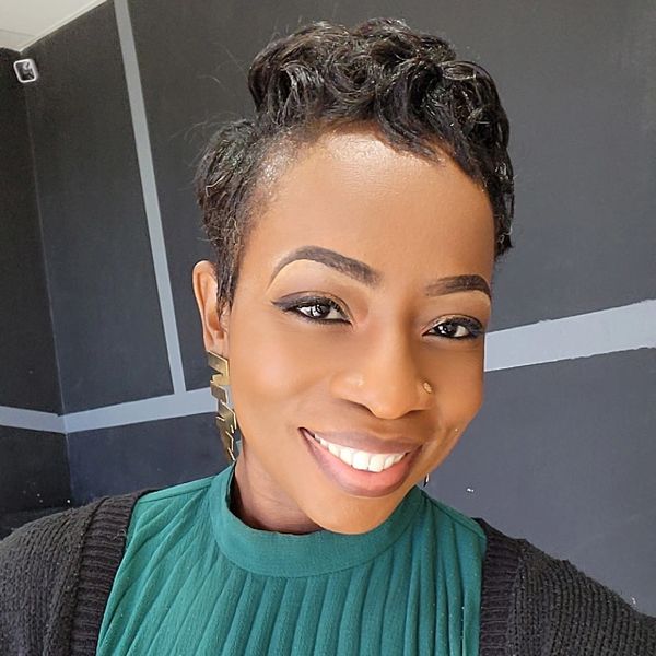 A woman with short curly hair smiles warmly, wearing a green top and black cardigan.