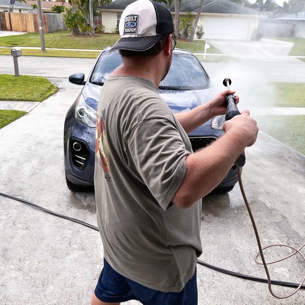 man wearing hat pressure washing car in a driveway