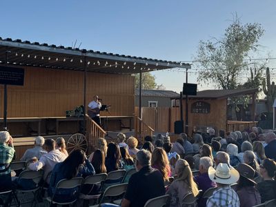 A man plays guitar on an outdoor stage to a seated audience at sunset.