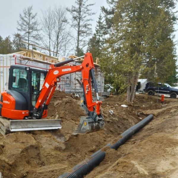 Orange excavator digging near pipes at a construction site with trees and buildings in the background.
