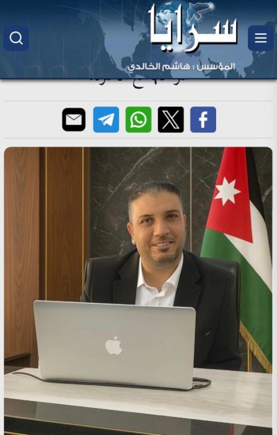 Man in suit sitting at desk with laptop and Jordanian flag behind.