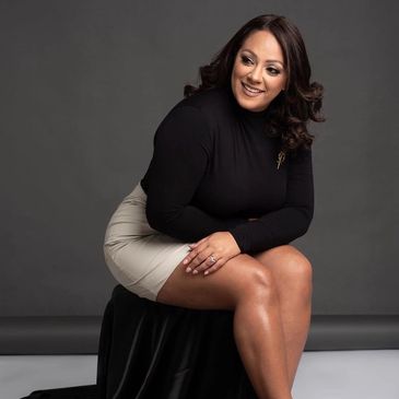 Woman in black top and beige skirt sitting on a stool against a dark background.