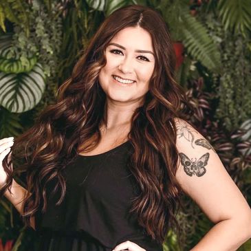 Smiling woman with long wavy hair and butterfly tattoo stands against leafy green backdrop.