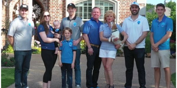 Group of eight people standing outdoors in front of a house, some holding a baby and a dog.