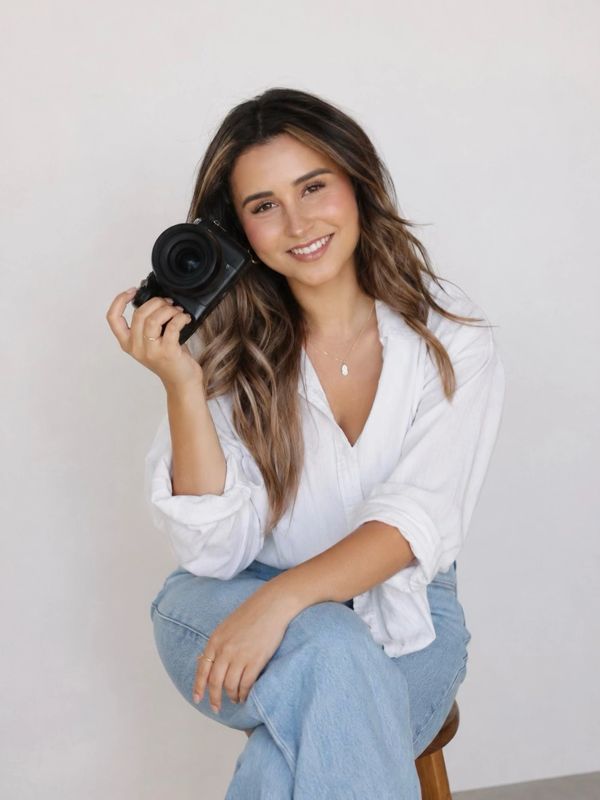 Smiling woman holding a camera, dressed casually in white and denim.
