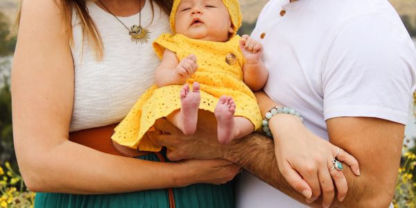 Parents holding a baby dressed in yellow outdoors.