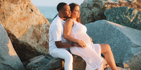 Couple embracing by rocky shore during sunset, expecting a baby.