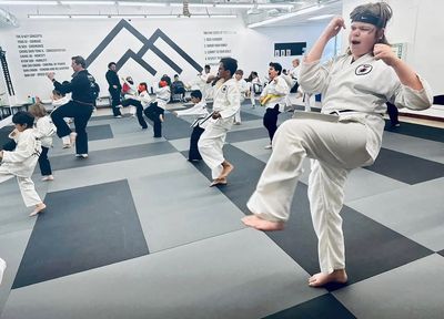 Kids and instructors practicing karate in a brightly lit dojo with padded gray floors.
