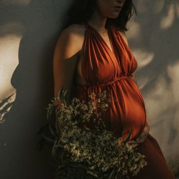 Pregnant woman in a rust-colored dress holding flowers, bathed in soft natural light.
