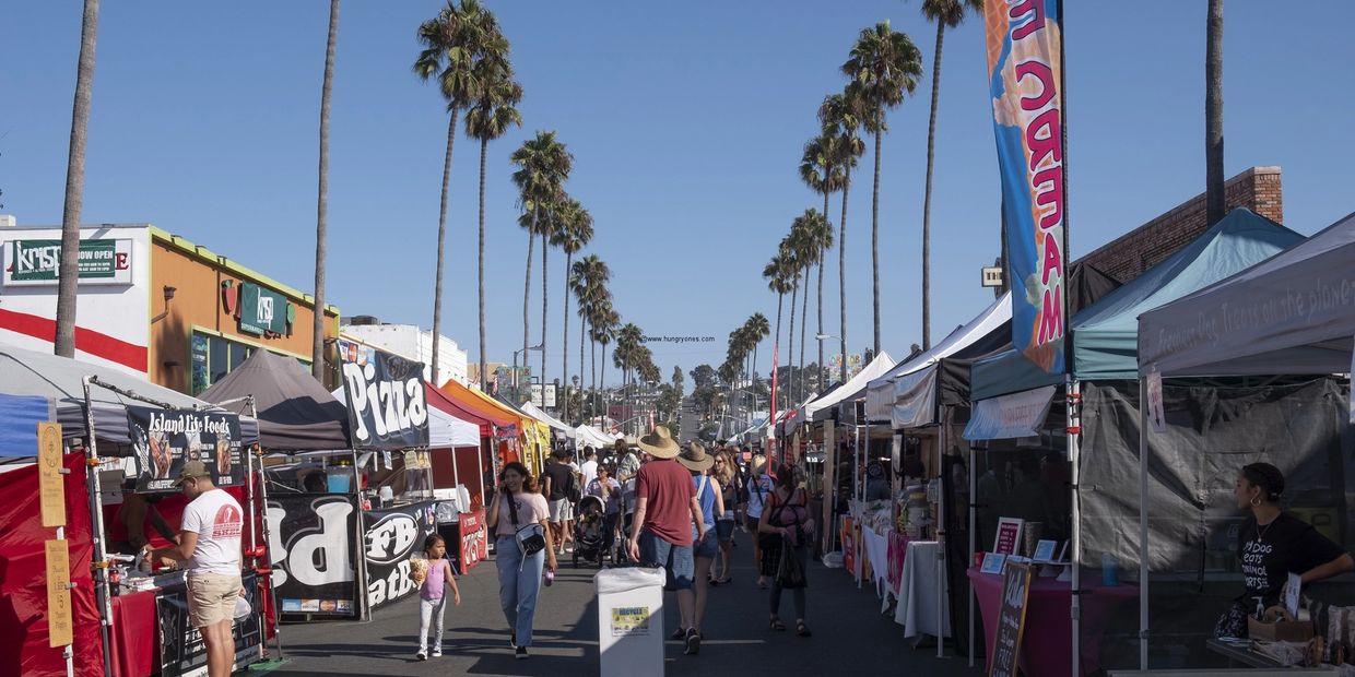 Outdoor market street with palm trees and various vendor tents under clear blue sky.