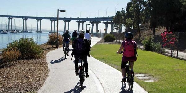 Group of cyclists riding on a waterfront path near a bridge on a sunny day.