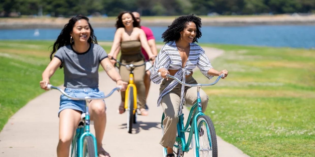 Four friends joyfully ride bikes on a sunny path near water.