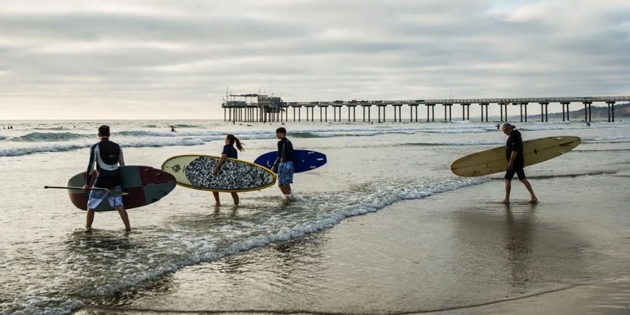 Four surfers entering the ocean with their boards near a pier.