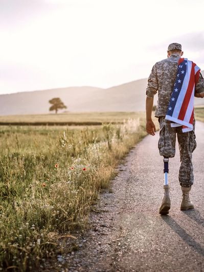 A soldier with a prosthetic leg walks down a rural road, carrying an American flag.