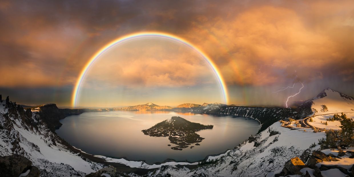 A vibrant double rainbow and lightning over a snowy mountainous lake at sunset.
