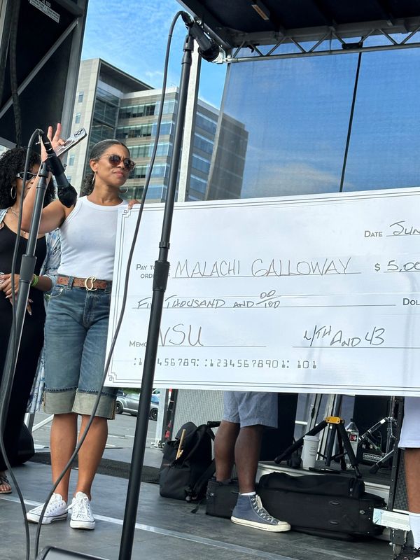 Woman in sunglasses holding a large check on stage during an outdoor event.