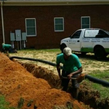 Worker digging a trench near a utility truck outside a brick building.